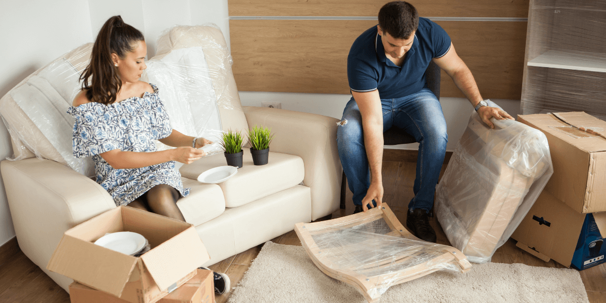 A man and woman are lifting and moving boxes in a spacious living room, preparing for a move or organizing their space.